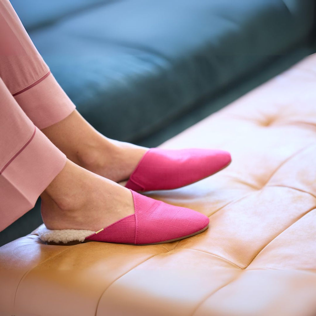 Woman wearing raspberry Luvons Devon slippers with shearling lining, sitting comfortably on a tan leather ottoman—stylish, supportive, and machine-washable.